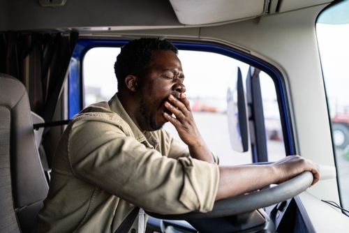 Truck driver yawning behind steering wheel inside cab, depicting fatigued driving condition